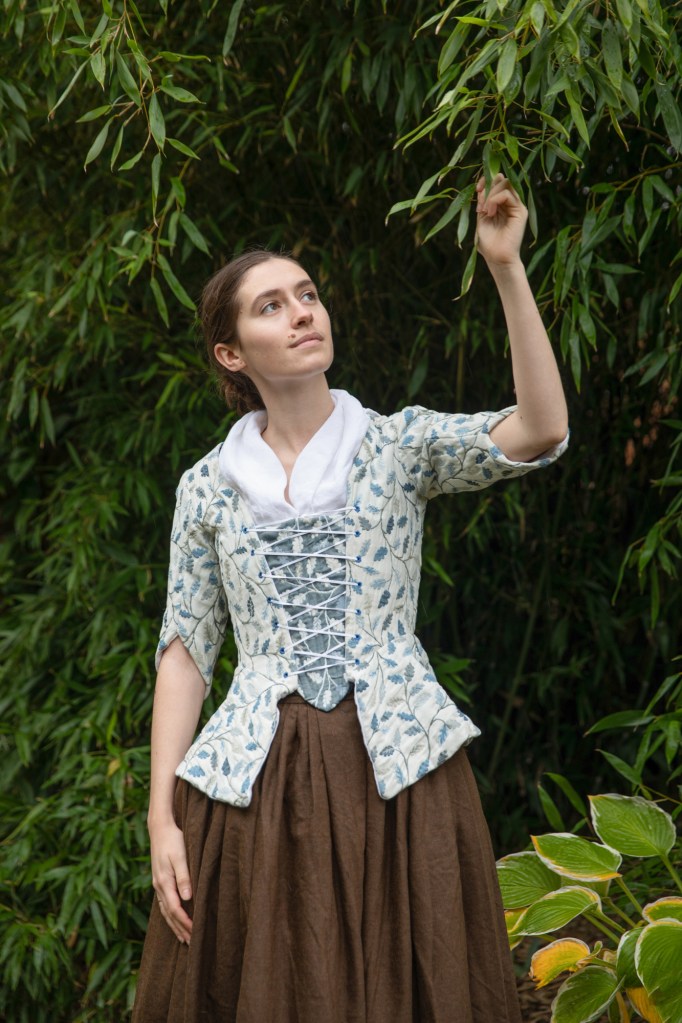 Katie looks up at a bamboo bush, wearing an 18th century outfit: a white jacket with blue leaf print and a brown wool skirt.