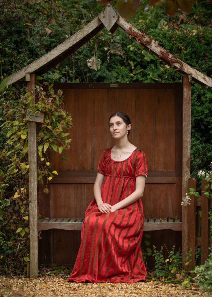 Katie sits thoughtfully in a red striped Regency dress on a garden bench